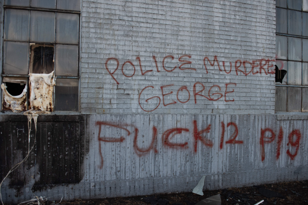 Ein Gebäude mit Graffiti an der Seite, das "Polizei Mord George Fuck 12 P19" lautet, mit Fenstern und diversen Gegenständen davor.