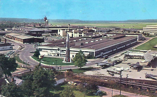 Altes Schwarz-Weiß-Foto eines Flughafens mit parkenden Flugzeugen, Gebäuden, Bäumen, Straßen, Fahrzeugen, Strommasten, Gras, Bahnschienen, Türmen, Hügeln und einem klaren blauen Himmel mit Text unten.