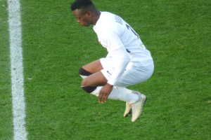 Ein professioneller Fußballspieler in weißer Uniform kniet auf einem Fußballfeld, trägt Fußballschuhe.
