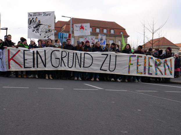 Demonstranten halten ein Transparent mit der Aufschrift "Kein Grund zu Feiern" gegen deutsche Sparmaßnahmen, mit Straßeninfrastruktur und Gebäuden im Hintergrund.