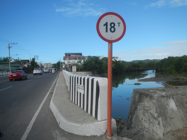 Ein Tempolimitschild am Straßenrand neben einem Fluss, mit Fahrzeugen, einer Barriere, Bäumen, Gebäuden, Strommasten mit Drähten und einem bewölkten Himmel im Hintergrund.