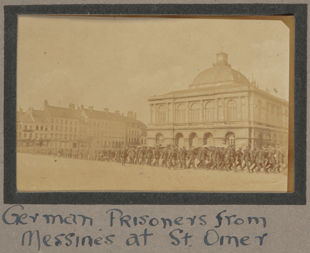Eine Gruppe deutscher Gefangener aus Messines steht vor einem Gebäude unter einem klaren blauen Himmel, mit Text am unteren Rand des Bildes.