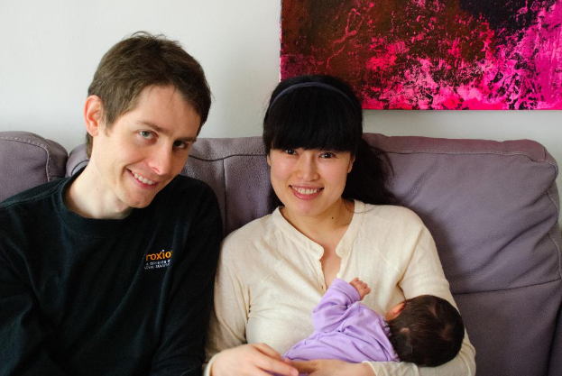 Eine Frau in einem weißen Hemd hält ein Baby und lächelt, während sie auf einem Sofa sitzt, neben einem Mann in einem schwarzen T-Shirt, der ebenfalls lächelt, mit einem rosafarbenen Fotoраhmen an der weißen Wand dahinter.