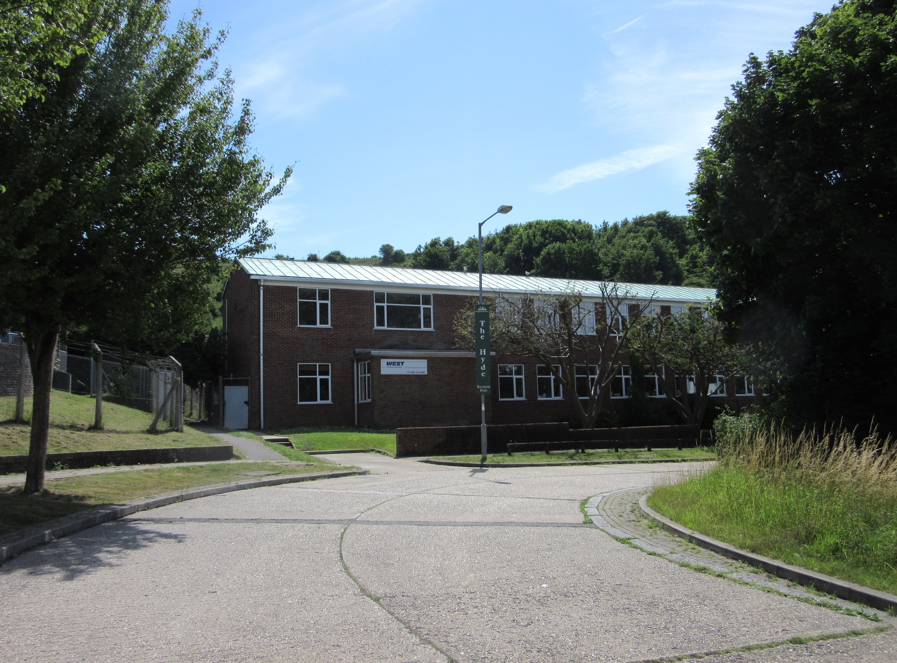 Ein zweistöckiges Schulgebäude mit mehreren Fenstern, einem Straßenmast, einem Schild und einer Gruppe von Bäumen, umgeben von saftigem Gras und einem Zaun, unter einem bewölkten Himmel.
