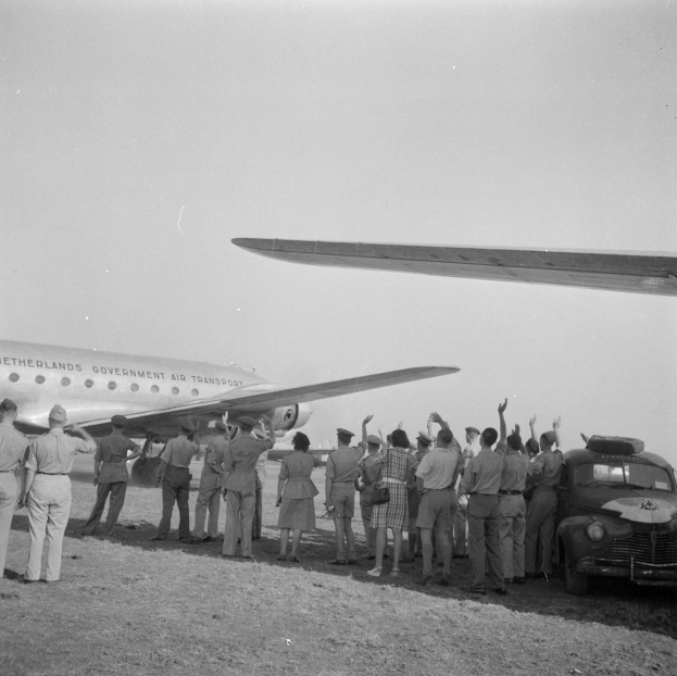 Eine Gruppe von Menschen, die um ein Flugzeug auf dem Boden stehen, mit einem parkenden Auto in der Nähe, dargestellt in Schwarz-Weiß.