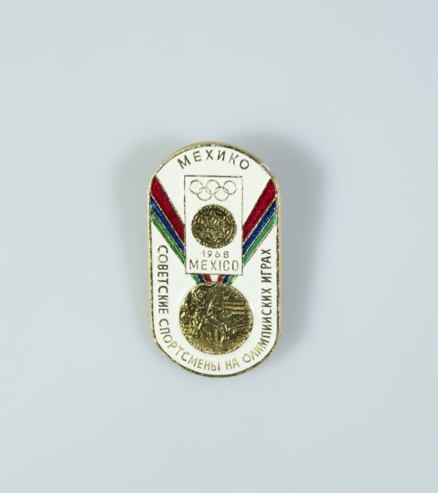 Ein Abzeichen mit der Aufschrift "1968 Mexico Olympic Games" und einer goldenen Medaille mit einer rot-weiß-blauen Schleife auf einem weißen Hintergrund.
