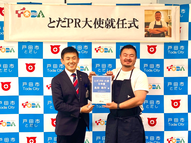 Zwei lachende Männer mit einem Zertifikat, im Hintergrund ein Banner mit der Aufschrift "Toda City Wins PR Award" und einem Foto einer Person.