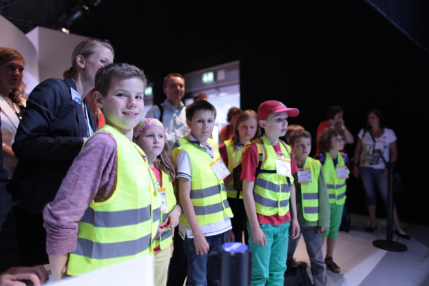 Eine Gruppe von Kindern mit Sicherheitswesten und Namensschildern, mit einem Jungen auf der linken Seite, der lächelt, auf einem Boden mit einem Pfahl auf der rechten Seite und ein paar Menschen im Hintergrund.