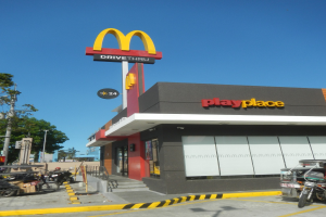 McDonald's-Restaurant mit einem "Playplace"-Schild, Fahrzeugen auf der Straße, Bäumen, Strommasten und einem klaren blauen Himmel im Hintergrund.