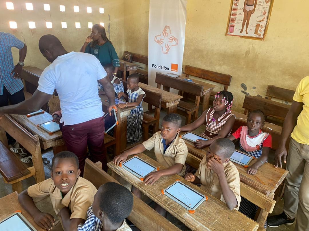 Kinder an Tischen sitzend, die Tablets in einem Klassenzimmer mit Erwachsenen in der Nähe, einem Banner und einem Plakat an der Wand verwenden.
