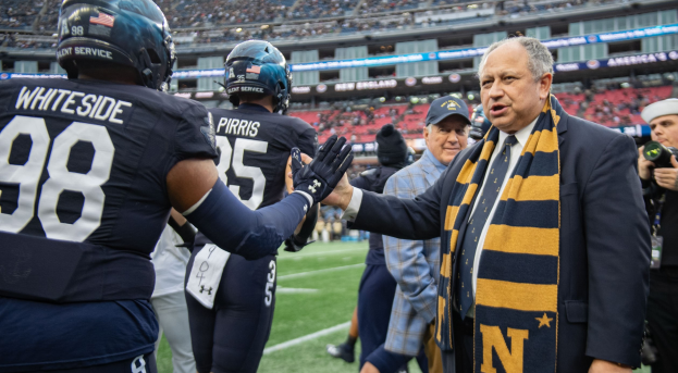 Ein Mann im Anzug schüttelt einem Fußballspieler die Hand auf einem Feld, mit einem Fotografen im Vordergrund und einem Stadion voller Zuschauer im Hintergrund.
