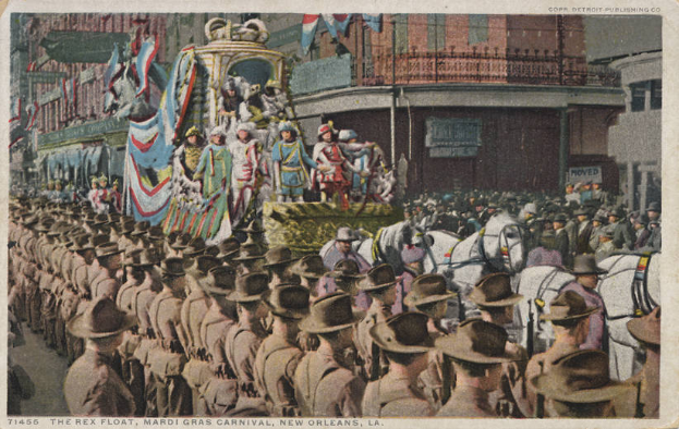 Eine alte Postkarte zeigt einen Mardi Gras-Umzug in New Orleans mit einem mit Fahnen und anderen Dekorationen geschmückten Festwagen, Reiter und Zuschauer am Straßenrand sowie im Hintergrund Gebäude mit Fenstern und Geländern.