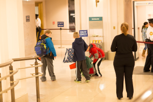 Eine Gruppe von Menschen steht in einem Flur mit einem Geländer auf der linken Seite, Deckenleuchten und Informationsschildern im Hintergrund, die den ersten Schultag im U.S. Capitol anzeigen.