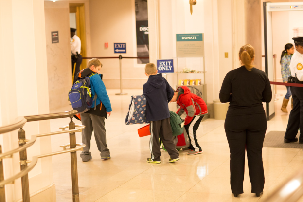 Eine Gruppe von Menschen steht in einem Flur mit einem Geländer auf der linken Seite, Deckenleuchten und Informationsschildern im Hintergrund, die den ersten Schultag im U.S. Capitol anzeigen.