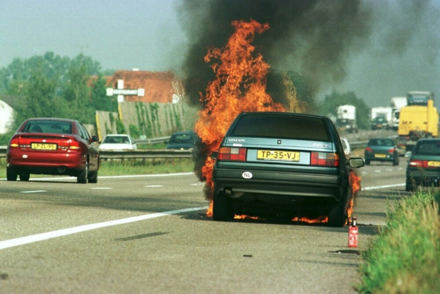 Ein brennendes Auto am Straßenrand mit Einsatzkräften in der Nähe, umgeben von anderen Fahrzeugen, Bäumen, Gebäuden und einem klaren blauen Himmel, mit Gras und einem Feuerlöscher auf der rechten Seite.