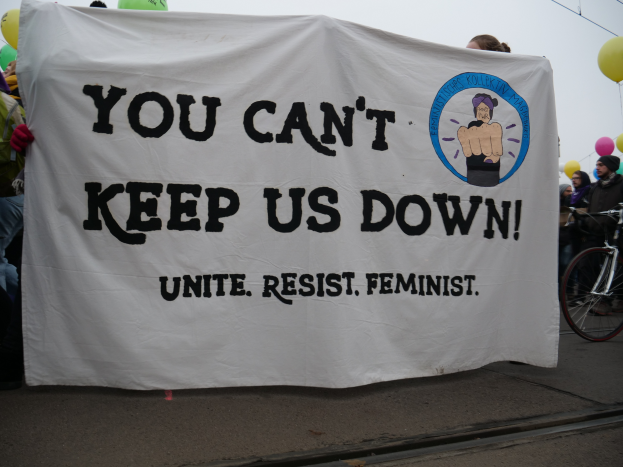 Eine Gruppe von Menschen steht auf der Straße und hält ein Banner in der Hand, auf dem "You Can't Keep Us Down, Unite Resist Feminist" steht, mit einem Fahrrad in der Nähe und Ballons, Drähten und einem klaren blauen Himmel im Hintergrund.