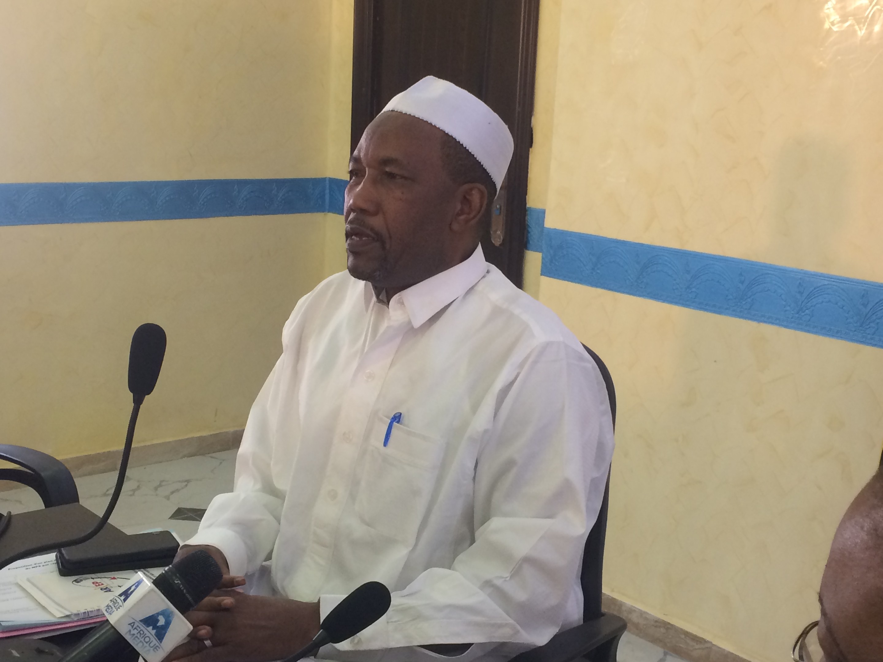 Ein Mann in einem weißen Gewand und einer Mütze sitzt an einem Tisch mit Mikrofonen, während er spricht und Papiere und Gegenstände auf dem Tisch sowie eine weitere Person mit Brille zu seiner Rechten zu sehen sind, in einem Raum mit einer Wand und einer Tür im Hintergrund.