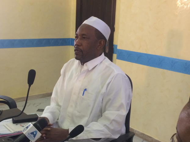 Ein Mann in einem weißen Gewand und einer Mütze sitzt an einem Tisch mit Mikrofonen, während er spricht und Papiere und Gegenstände auf dem Tisch sowie eine weitere Person mit Brille zu seiner Rechten zu sehen sind, in einem Raum mit einer Wand und einer Tür im Hintergrund.