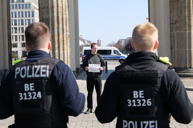 Drei Polizeibeamte stehen vor einem Gebäude mit einem Mann, der ein Papier hält, flankiert von Säulen, während Fahrzeuge, Menschen, Bäume und Gebäude den Hintergrund und den Himmel ausfüllen.