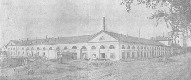 Schwarz-weiß-Foto eines großen, mehrfenstrigen Gebäudes, das von Grün umgeben ist und unter einem klaren Himmel steht, mit dem Text "1892 – das erste Fabrikgebäude der Stadt" unten.