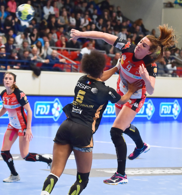 Eine Gruppe von Frauen, die auf einem Handballfeld mit einem Ball in der Luft spielen, beobachtet von Zuschauern in einer Stadionatmosphäre.