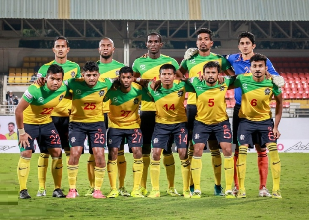 Eine Gruppe von Männern auf einem Fussballfeld, die einen Sieg feiern, umgeben von Werbetafeln, Stühlen, Geländern, Pfosten, Bäumen und einer Hütte im Hintergrund.