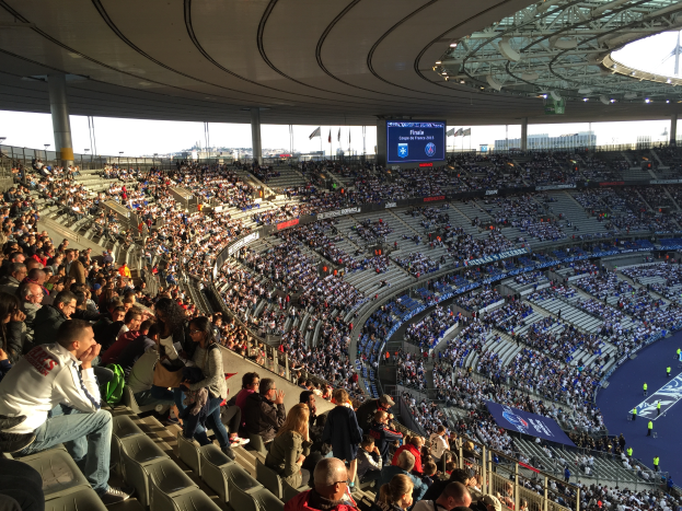 Eine große Menschenmenge sitzt im Stadion und schaut ein Fußballspiel, mit einer Bühne auf der rechten Seite, Fahnen, Stangen und einem Bildschirm im Hintergrund, unter einem sichtbaren Himmel, im Allianz Arena in München, Deutschland.