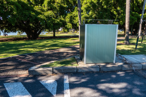 Tragbare Toilette am Straßenrand in einem Park mit Bäumen, Gras, einem Gewässer und einem klaren blauen Himmel im Hintergrund.