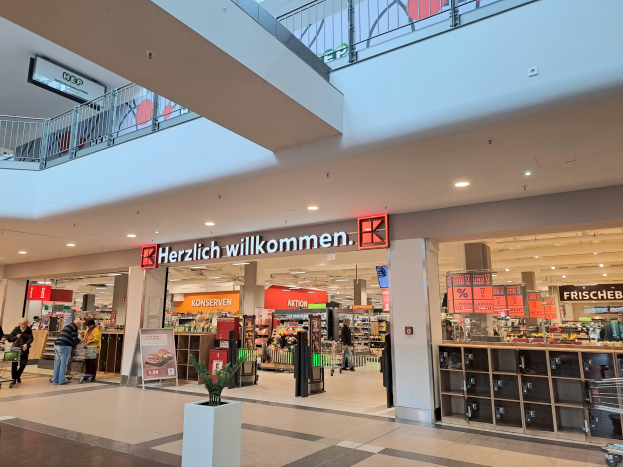 Das Innere eines Einkaufszentrums mit Menschen, die herumlaufen, Geschäften, Schildern mit Text, Lampen, Geländern und einer Topfpflanze unten; der Text auf den Schildern lautet "Herzlich willkommen".