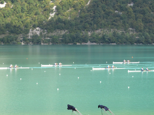 Eine Gruppe von Menschen, die auf einem Gewässer umgeben von Bäumen und einem Hügel rudern, mit Bekleidung-behangenen Tribünen unten, was auf einen Ruderwettbewerb hinweist.
