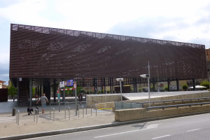 Großes Gebäude mit zahlreichen Fenstern, das als neuer Eingang zum Stadtzentrum dient, umgeben von Straßeninfrastruktur, Fahrzeugen, Fußgängern, Bäumen und einem bewölkten Himmel.