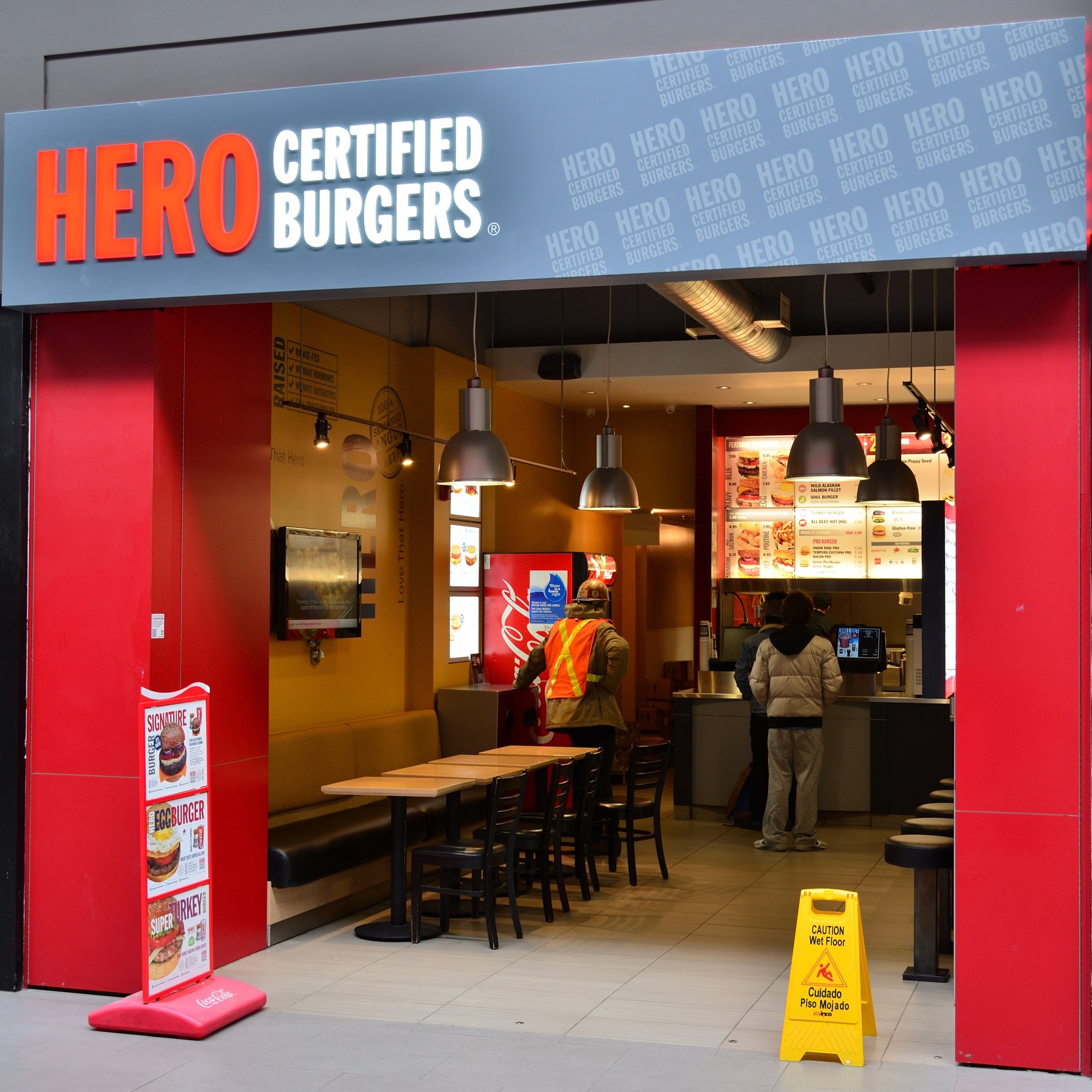 Außenansicht eines Hero Certified Burgers Restaurants in London, England, mit Gästen, Sitzgelegenheiten, einer Speisekarte, einem Wandfernseher und Deckenleuchten.