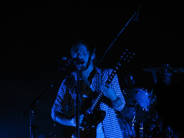 Ein Mann in der Mitte des Bildes spielt eine Gitarre und singt in ein Mikrofon, während hinter ihm in einem dunklen Konzertsetting Trommeln zu sehen sind.
