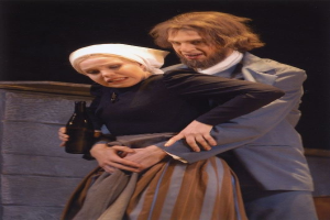 Ein Mann in einem Anzug mit ernster Miene und eine Frau mit einer Flasche stehen auf einer Bühne mit Text unten und einer Wand im Hintergrund.