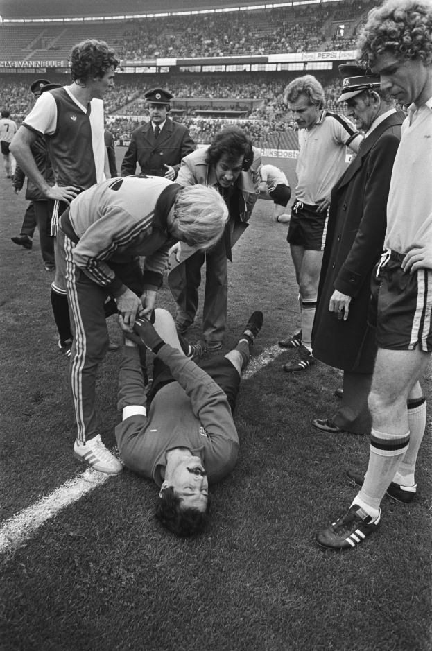 Schwarzes und weißes Foto eines Fußballspielers, der auf dem Boden liegt und von einer Gruppe Männer umgeben ist, mit einer Menge im Stadionhintergrund.
