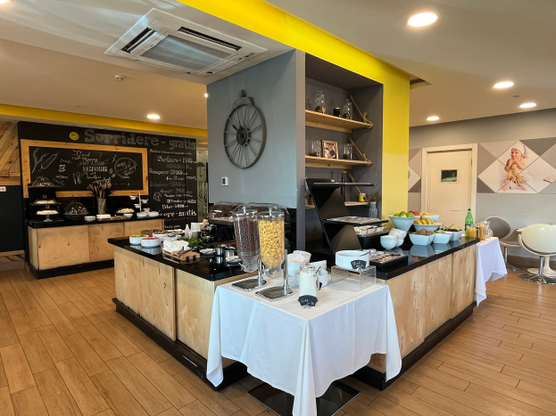 Ein Restaurant-Innenraum mit Tischen und Stühlen, Lebensmitteln, einer Tafel mit Text, einer Wanduhr, Deckenleuchten, einer Klimaanlage und einer Tür im Hintergrund, Located im Hôtel de Ville in Paris.