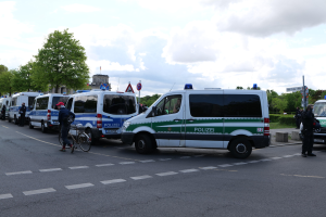 Eine Gruppe von Polizeiwagen, die entlang einer Straße geparkt sind, mit einem Radfahrer im Vordergrund und Gebäuden, Bäumen, Schildern und einer bewölkten Himmel im Hintergrund.