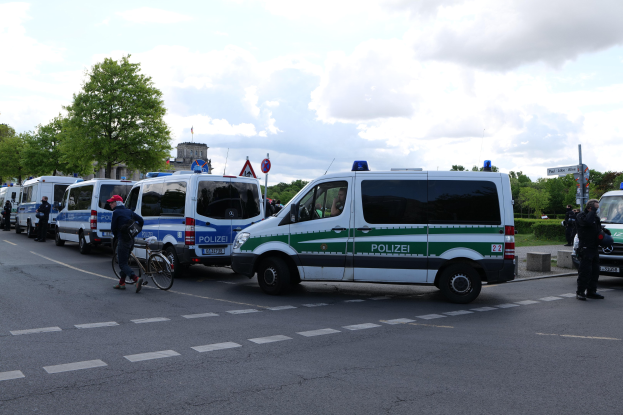 Eine Gruppe von Polizeiwagen, die entlang einer Straße geparkt sind, mit einem Radfahrer im Vordergrund und Gebäuden, Bäumen, Schildern und einer bewölkten Himmel im Hintergrund.
