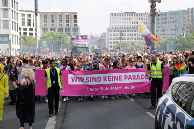 Eine große Gruppe von Menschen marschiert die Straße entlang und hält ein 'Happy Pride March Berlin'-Schild, mit einem geparkten Auto auf der rechten Seite und Gebäuden, Bäumen, Pfählen und Lichtern im Hintergrund unter einem klaren blauen Himmel.