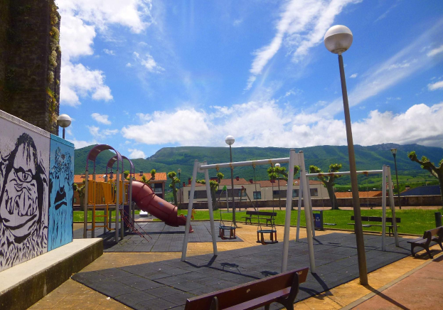 Spielplatz mit Schaukeln, einer Rutsche, Bänken, Pfosten, Lichtern, einer bemalten Wand, Gras, Bäumen, entfernter Gebäude, Hüllen und einem bewölktem Himmel.