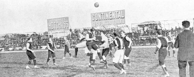 Ein Schwarz-Weiß-Foto von einer Gruppe von Männern, die auf einem Feld Fußball spielen, mit einem Ball in der Luft und Zuschauern im Hintergrund, mit Texttafeln, die wahrscheinlich Matchdetails angeben.