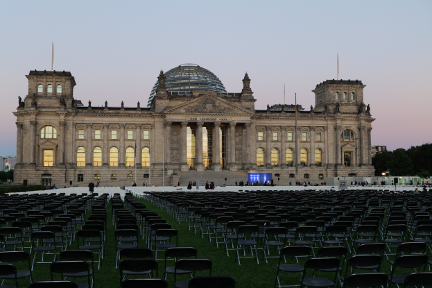 Das Reichstagsgebäude in Berlin, Deutschland, mit seinen Fenstern, Säulen, Bögen und Kuppel, umgeben von Menschen, Stühlen auf dem Rasen, Straßenlaternen, Schildern, Bäumen und einem bewölkten Himmel.