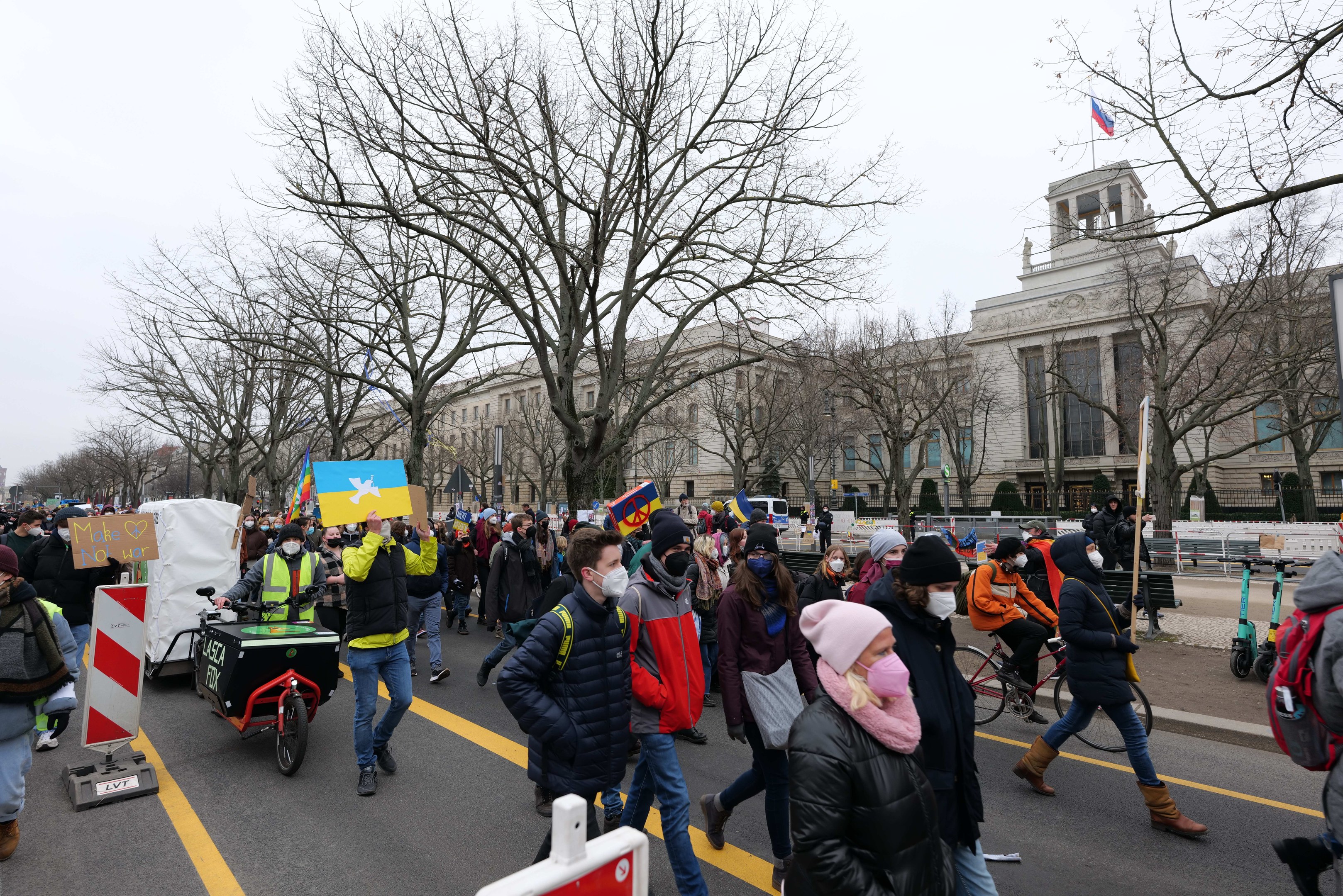 Eine große Gruppe von Menschen marschiert auf einer Stadtstraße in einem Protest, einige halten Schilder und andere fahren Fahrräder, mit Bäumen und Gebäuden im Hintergrund unter einem klaren blauen Himmel.
