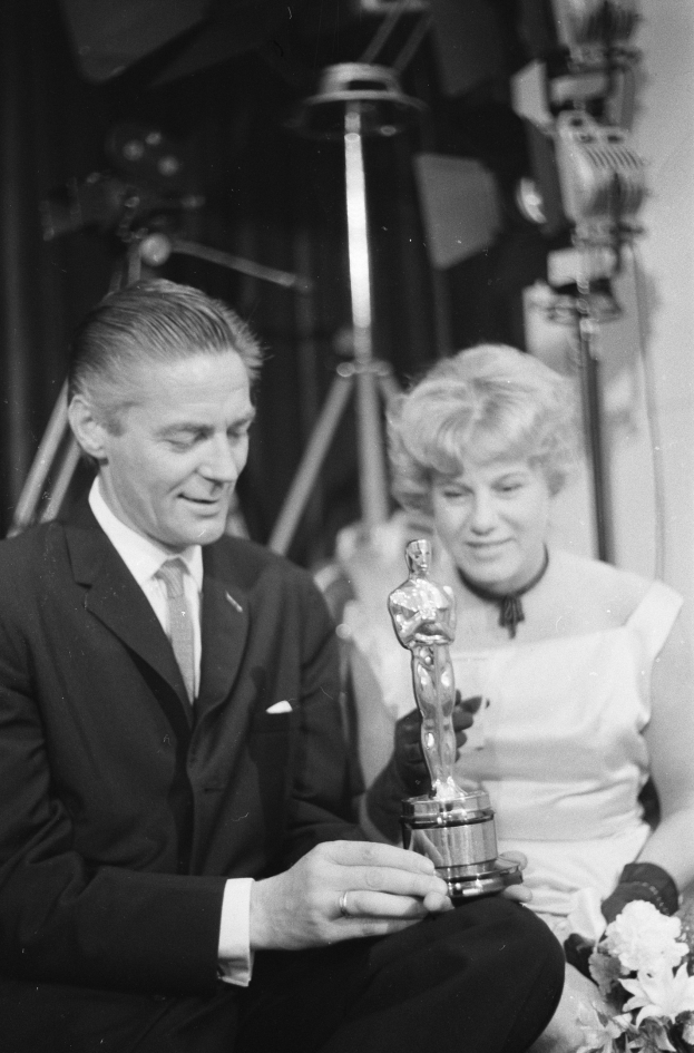 Schwarzes und weißes Bild eines Mannes in einem schwarzen Blazer, weißem Hemd und Krawatte, der eine Oscar-Statue und eine Frau in Abendgarderobe mit einem Blumenstrauß in der Hand hält, nebeneinander sitzend mit Hintergrundbeleuchtung.