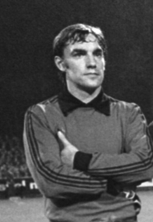 Ein Schwarz-Weiß-Foto eines älteren Mannes auf einem Fußballfeld mit verschränkten Armen, umgeben von einer Gruppe von Menschen im Hintergrund.