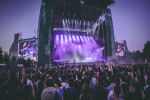 Eine große Menschenmenge bei einem Musikfestival schaut Künstlern auf einer hell erleuchteten Bühne mit Bildschirmen zu, die vor einem Gebäude, Bäumen und einem klaren blauen Himmel steht.