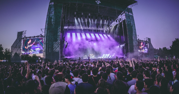 Eine große Menschenmenge bei einem Musikfestival schaut Künstlern auf einer hell erleuchteten Bühne mit Bildschirmen zu, die vor einem Gebäude, Bäumen und einem klaren blauen Himmel steht.