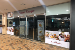 Der Eingang eines Restaurants namens "Geheimes Rezept" hat Glastüren, einen Stand mit verschiedenen Gegenständen und eine Speisekarte auf der linken Seite sowie warmes Deckenlicht über der Tür.