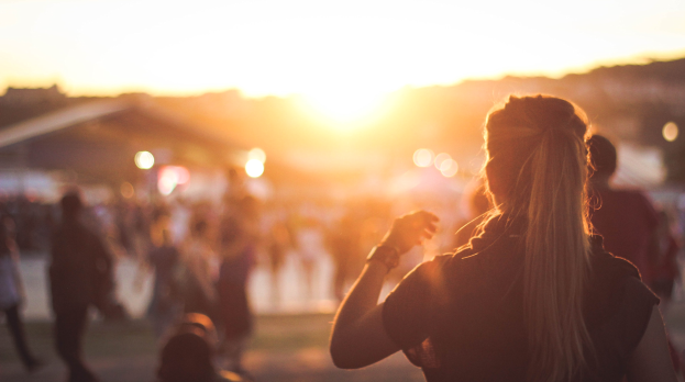 Eine Frau vor einer Menge bei einem Musikfestival bei Sonnenuntergang, mit einem orangefarbenen, pinken und violetten Himmel im Hintergrund.