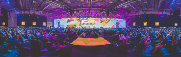 Eine große Menschenmenge sitzt auf Stühlen vor einer hell erleuchteten Bühne mit mehreren stehenden Personen, wahrscheinlich bei einem Online-Marketing-Rockstars-Event, umgeben von Bildschirmen.
